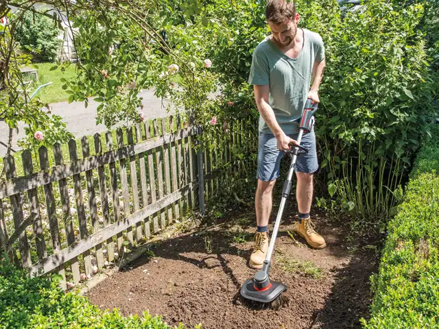 Mann mit Gartengerät auf Beet umgeben von Bäumen und Büschen