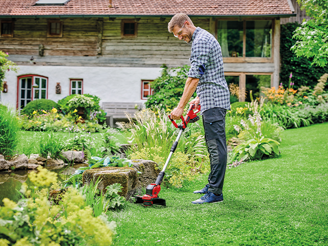 Mann trimmt mit Akku-Trimmer Rasen im Garten vor Teich