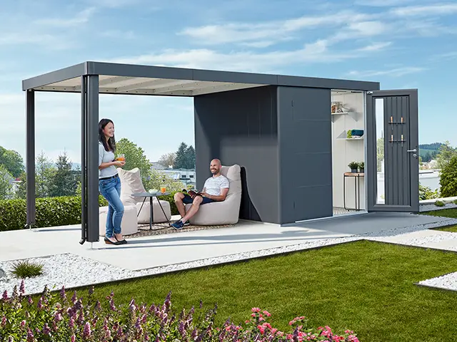 Ein Mann sitzt und eine Frau steht in einem modernen Gartenpavillon auf einer Terrasse.
