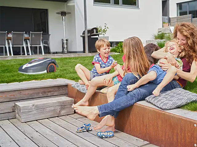 Kinder auf Terrasse - im Hintergrund mäht Rasenmähroboter Rasen