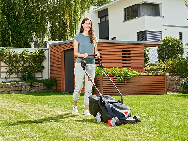 Frau mit Rasenmäher auf Rasenfläche vor modernem Gartenhaus