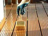 Eine Hand mit Handschuh streicht Holzlatten einer Terrasse im Freien mit einem Pinsel.