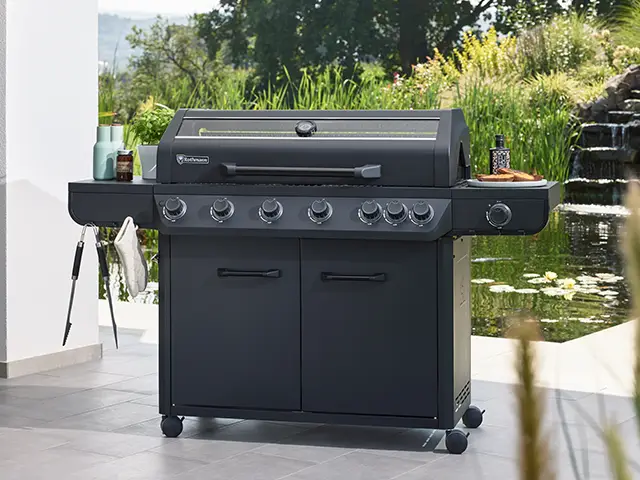 Ein großer Rothmann Grill steht auf einer Terrasse vor einem Teich mit Pflanzen im Hintergrund.