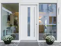 Eine weiße Tür mit Glaseinsatz, flankiert von Fenstern, steht zwischen zwei Blumentöpfen.