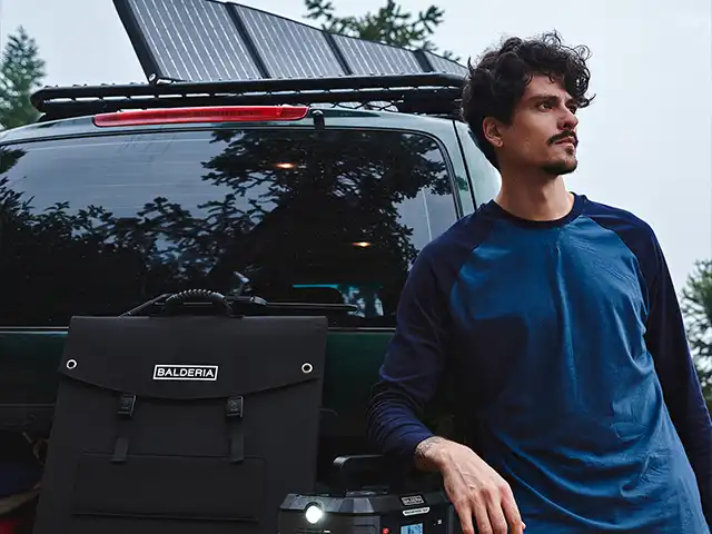 Ein Mann lehnt an einem Auto mit Solarpanelen, im Wald, Tasche mit "BALDERIA".