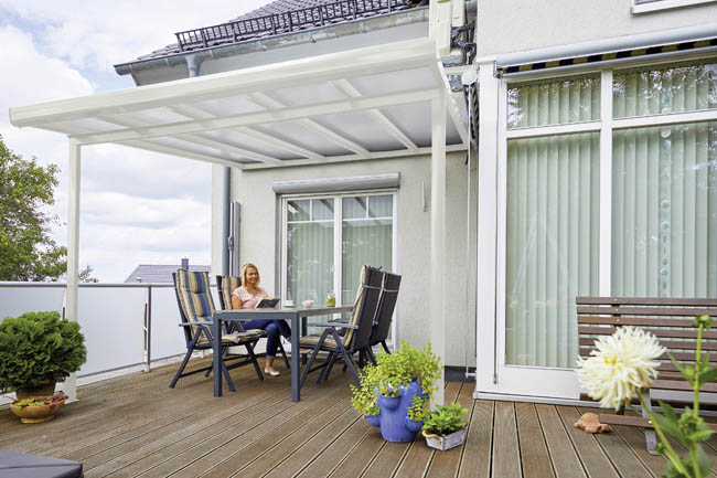 Frau sitzt unter einer weißen Terrassenüberdachung an einem Gartentisch auf einer Holzterrasse
